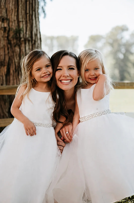 A smiling woman with two young girls wearing matching white sleeveless tulle dresses with pearl sash