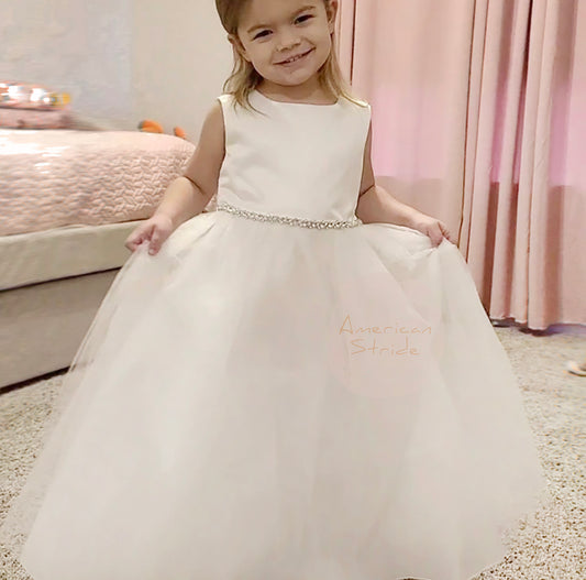 Young girl in a white dress with 'American Stride' branding, standing on a carpeted floor.
