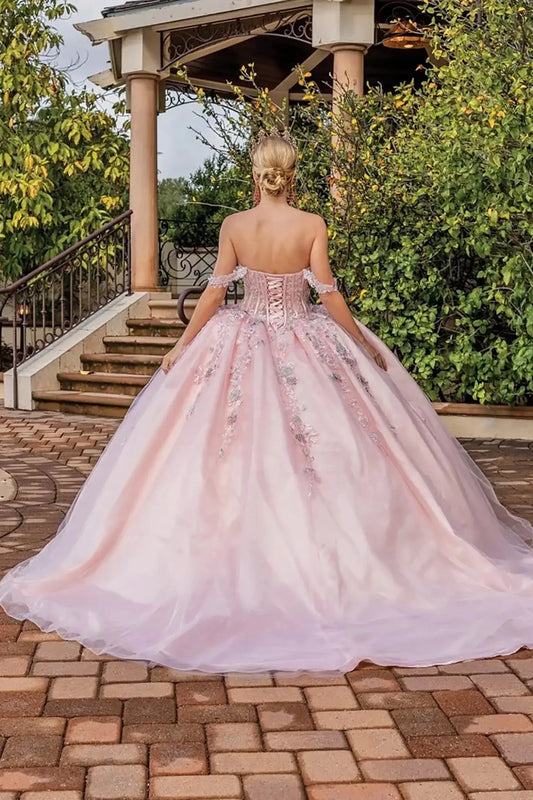 Back view of a blush pink off-shoulder quinceañera ball gown featuring a lace-up corset back, floral appliqués, flowing tulle skirt, and soft train.