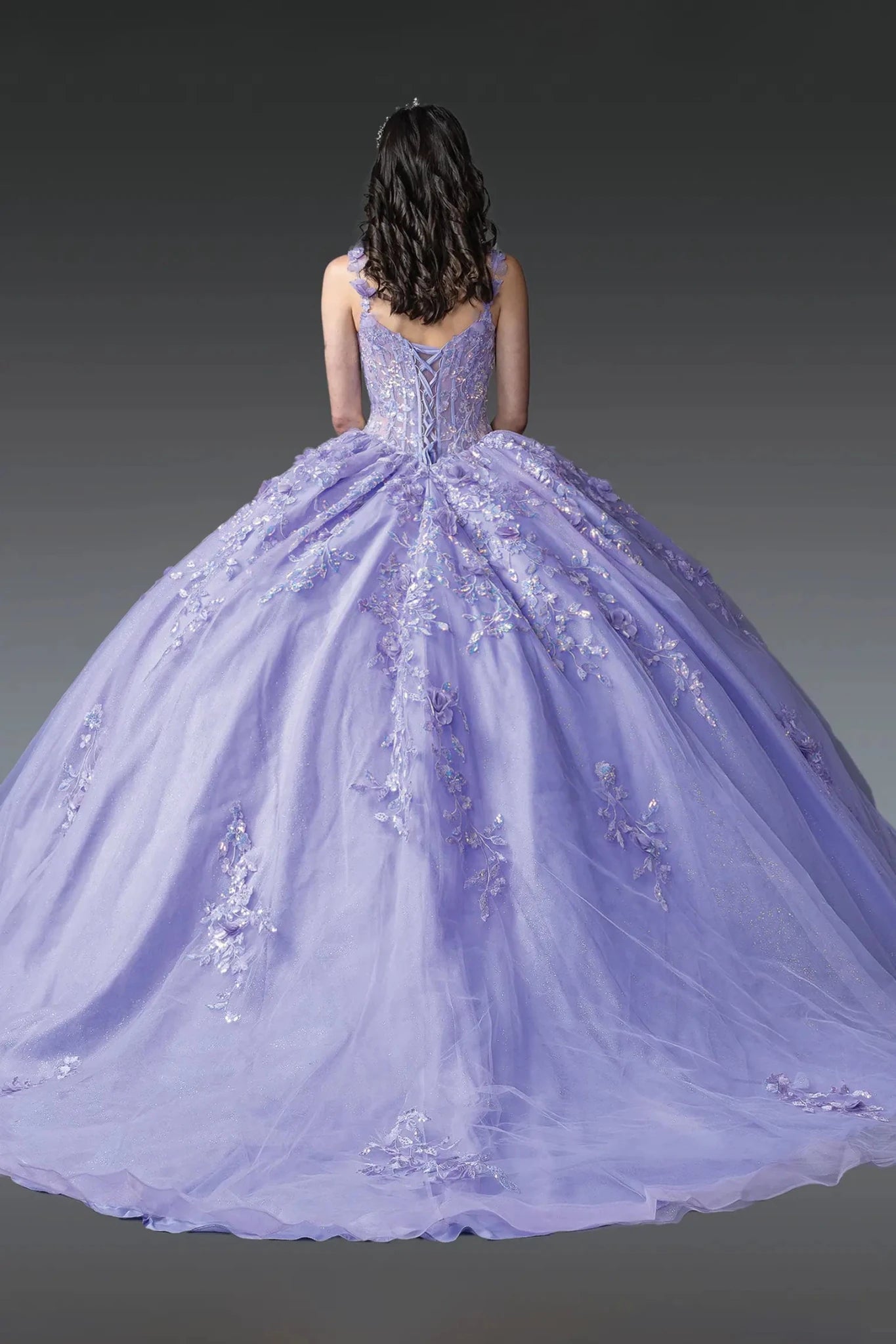 Woman standing back-facing in lilac quinceañera ball gown with lace-up bodice, embroidered floral details, sheer overlay, and flowing layered skirt with train