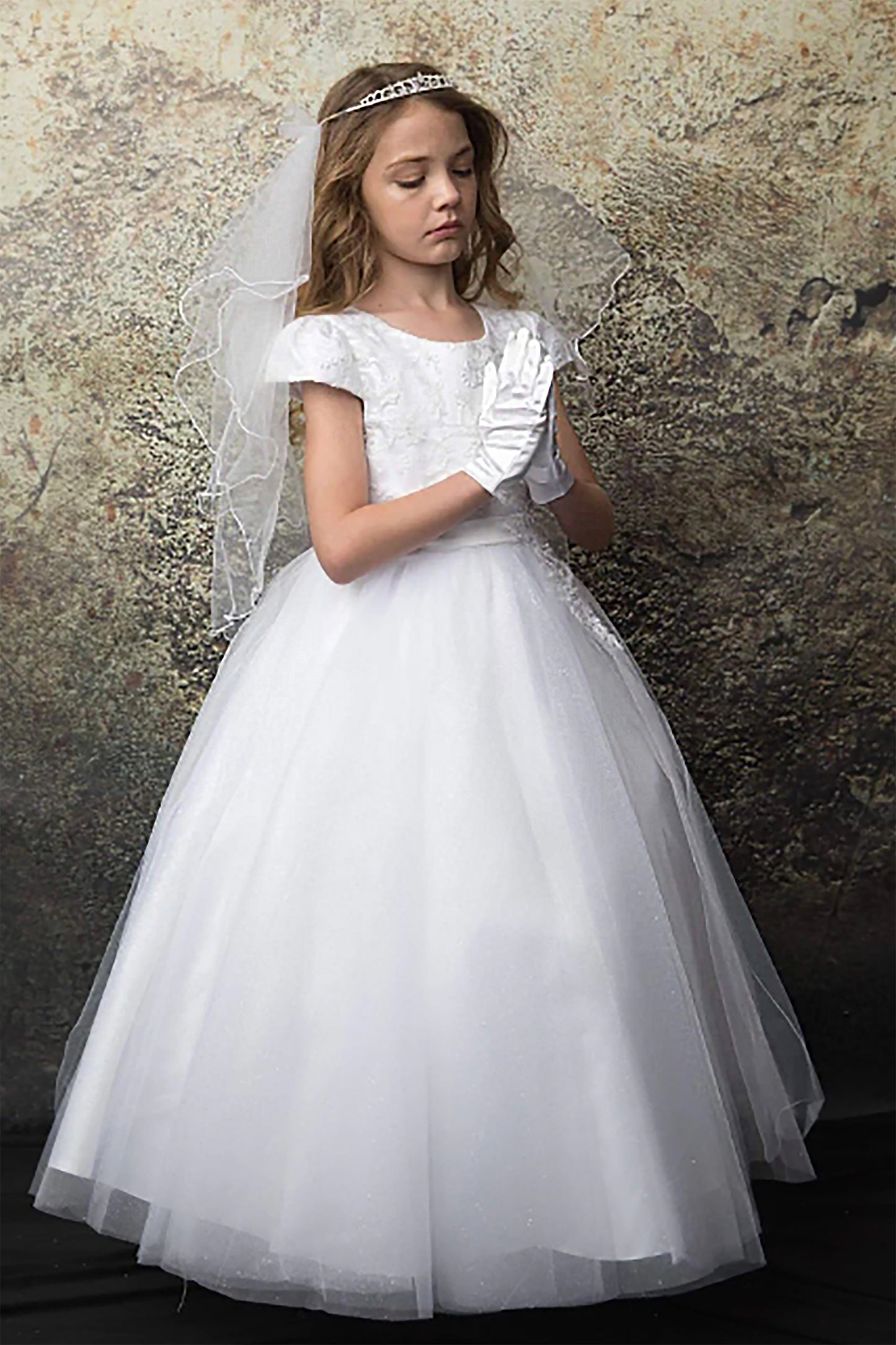 Young girl in a white formal dress with a veil against a textured wall.