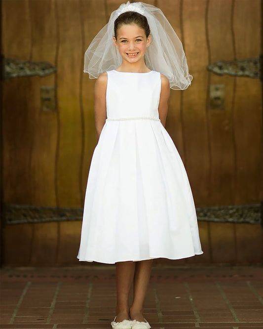 Young girl in a white dress with a veil standing against a wooden wall.