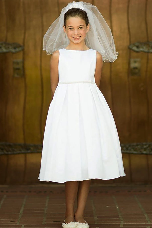 Young girl in a white dress and pearl/rhinestone belt with a veil standing against a wooden wall.