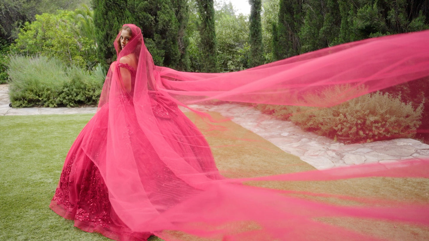 woman standing outdoors on a grassy area beside a stone pathway, dressed in a vibrant red traditional outfit. The attire includes a long, flowing veil that extends dramatically behind her, creating a striking visual effect.