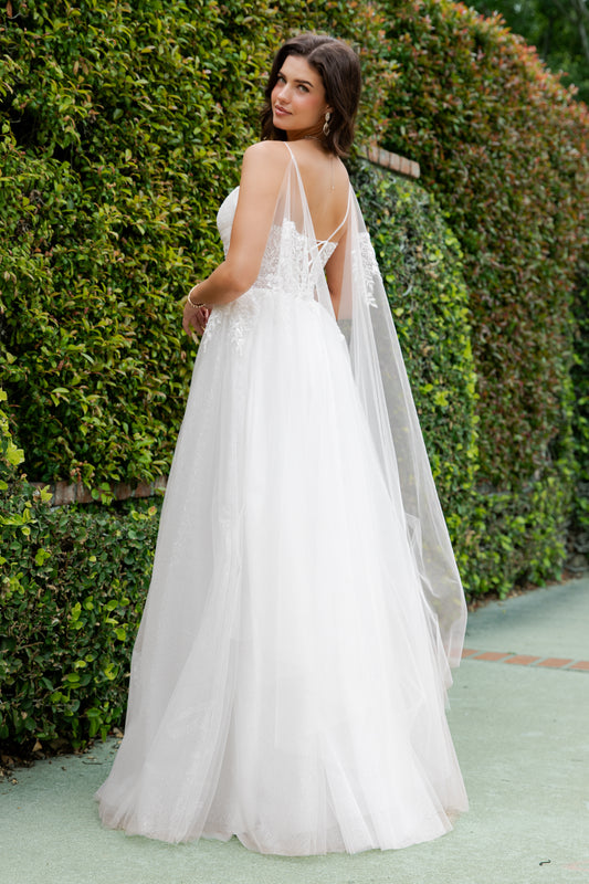 Woman in a white wedding dress with a cape standing in front of green bushes.