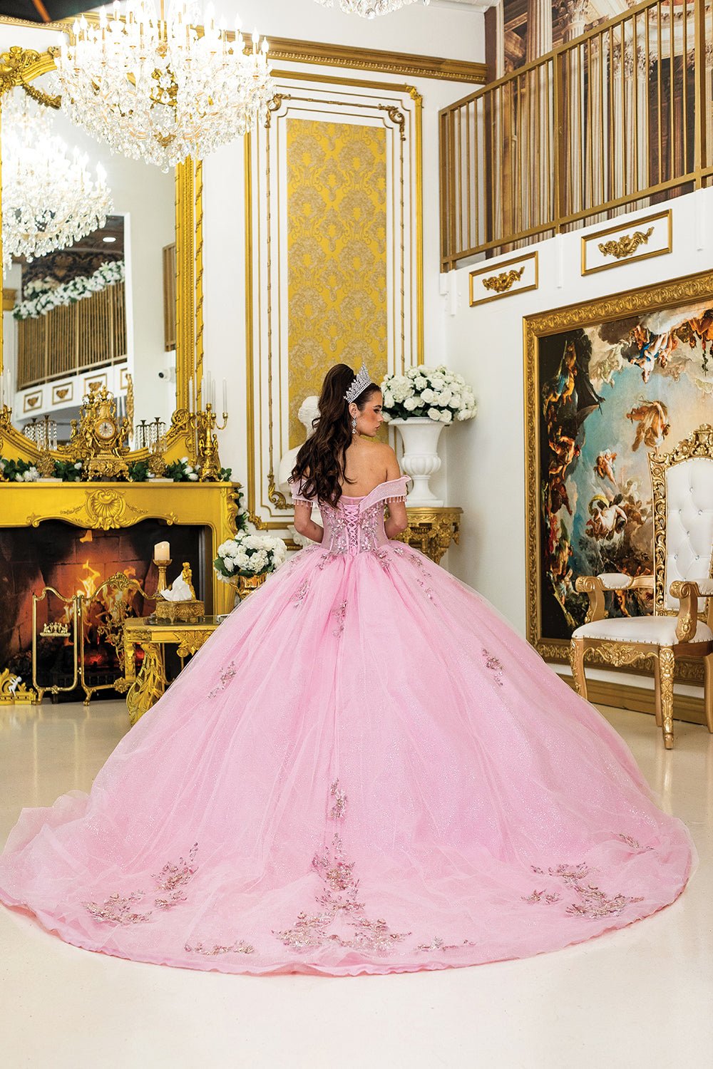 Woman standing back-facing in pink quinceañera ball gown with off-shoulder beaded fringe sleeves, lace-up sheer corset bodice, 3D floral appliqués, and flowing glitter tulle skirt with train