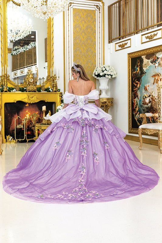 Woman standing back-facing in lilac-white quinceañera ball gown with lace-up embroidered bodice, floral embellishments, and flowing layered skirt with train