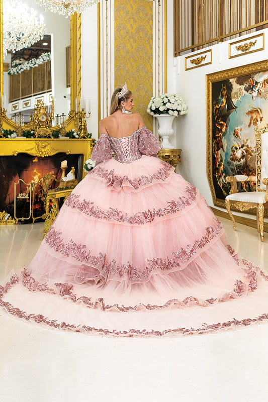 Woman standing back-facing in rose gold quinceañera ball gown with lace-up embroidered bodice, off-shoulder sleeves, glitter accents, and flowing layered skirt with train
