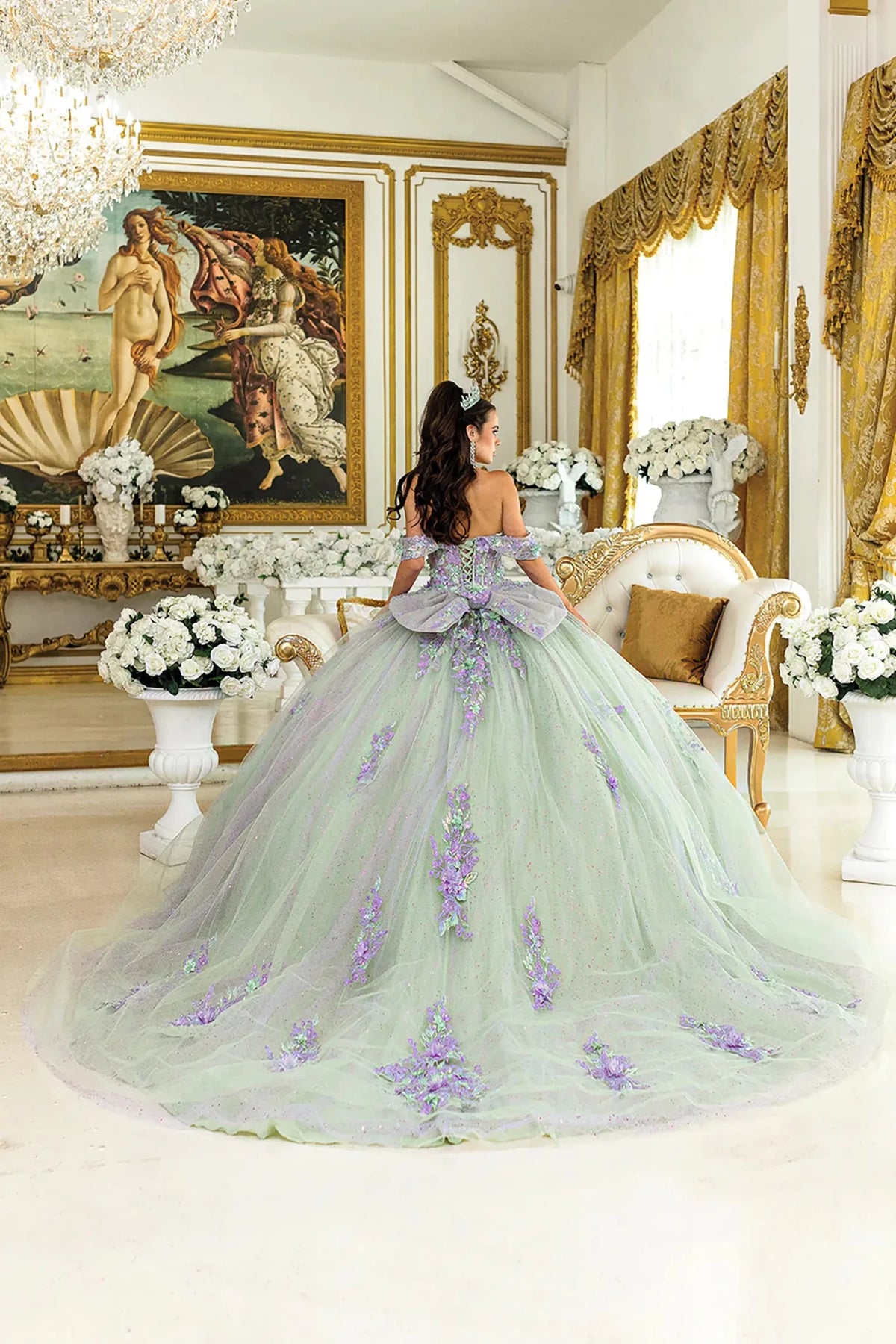 Woman standing front-facing in lilac-sage quinceañera ball gown with off-shoulder floral appliqués, corset bodice, glittered tulle skirt, and layered detailing