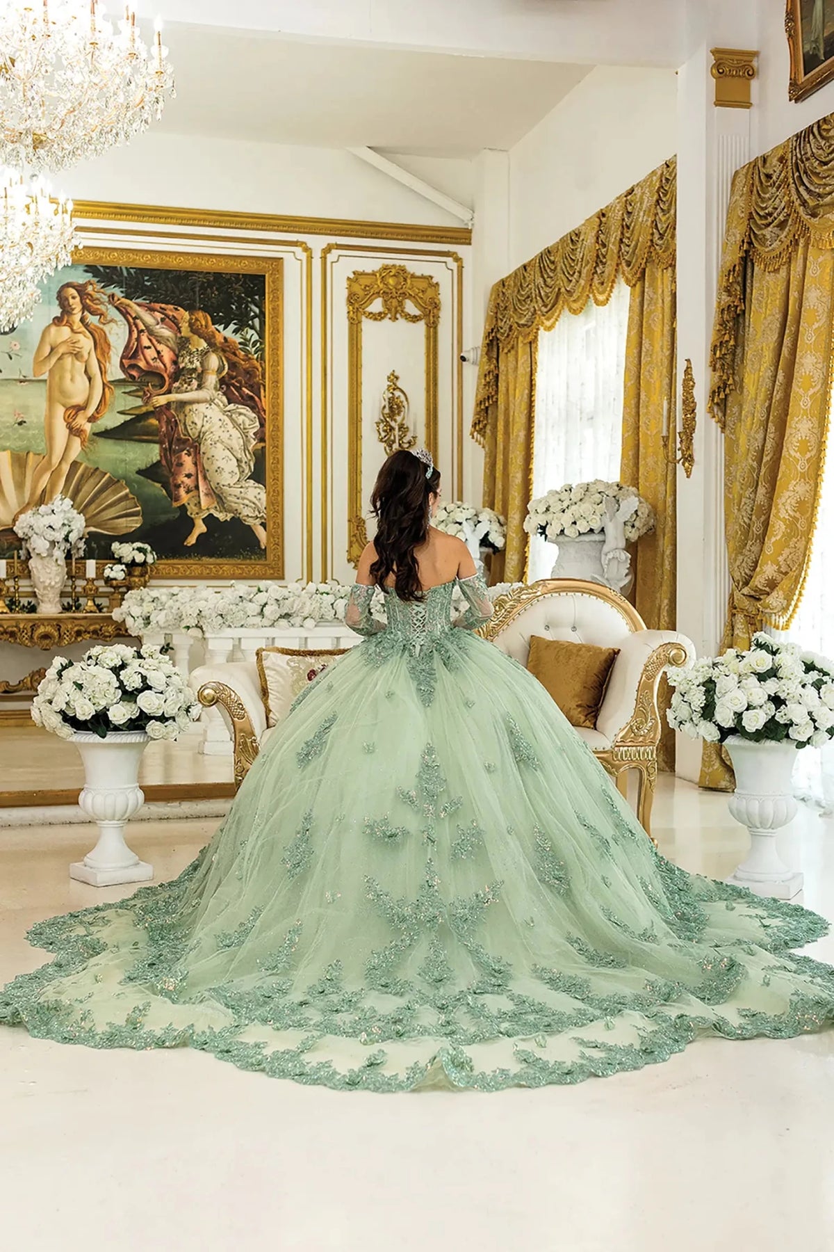 Woman standing back-facing in lilac-sage quinceañera ball gown with lace-up corset bodice, off-shoulder floral appliqués, glittered tulle skirt, and flowing layered train