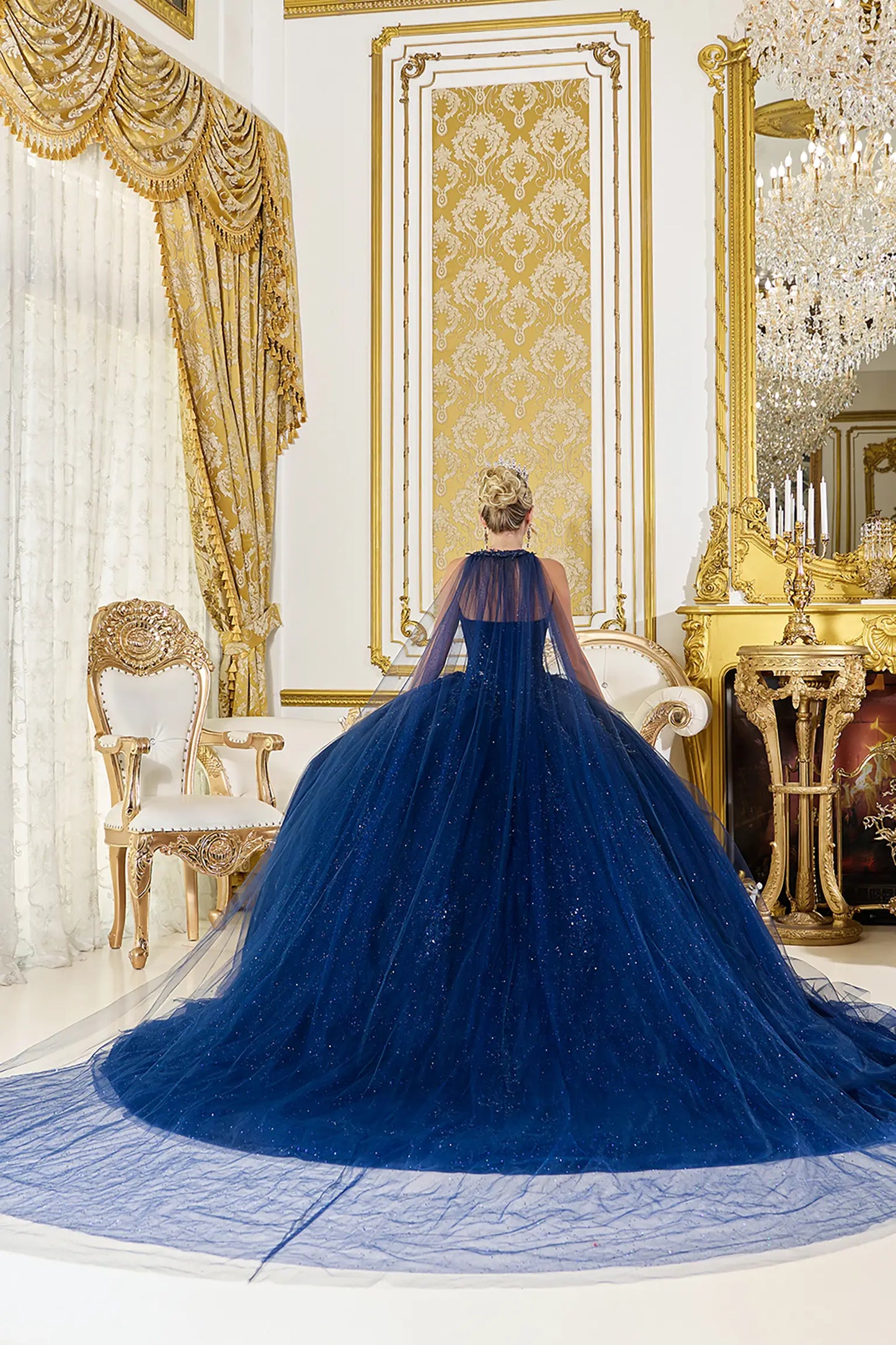 Back view: Navy blue quinceañera ball gown back view featuring a sheer cape-style overlay, flowing tulle skirt with subtle sparkle, and an extended train in an elegant ballroom interior