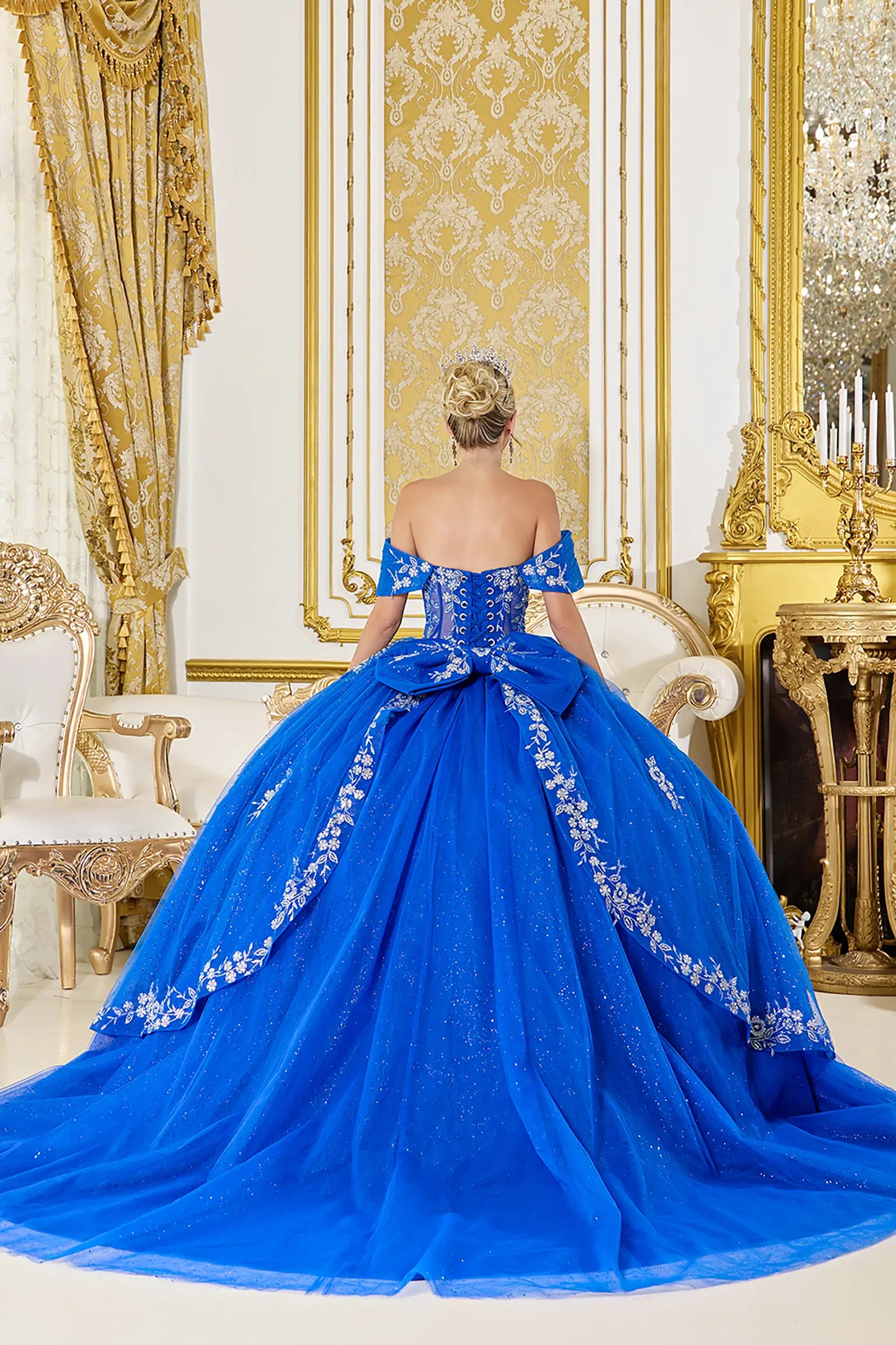 Back view of royal blue quinceañera ball gown showcasing lace-up corset back, oversized bow accent, embroidered skirt panels, and sparkling tulle train in a luxurious interior
