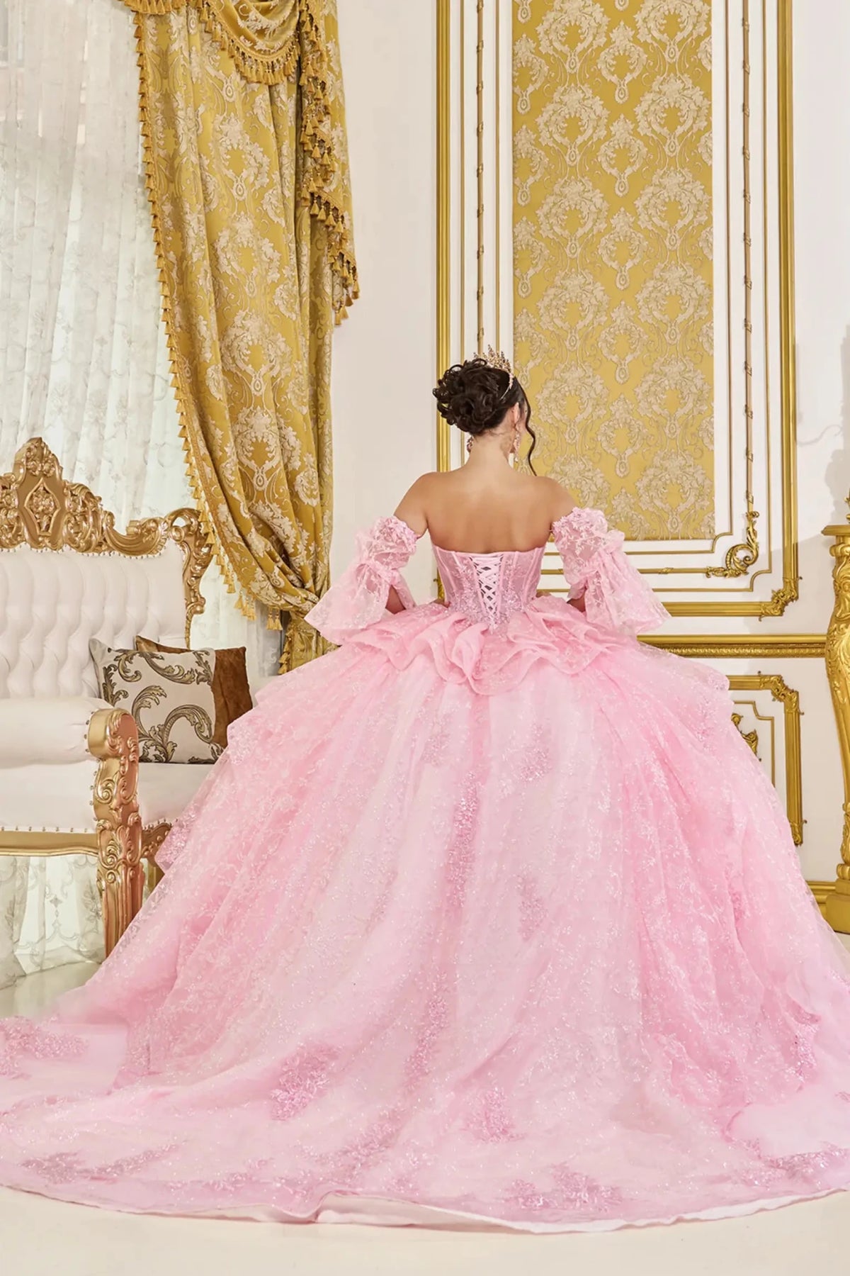 Back view of pink quinceañera ball gown featuring lace-up corset back, off-the-shoulder sleeves, and full layered glitter tulle skirt in an elegant ballroom setting.