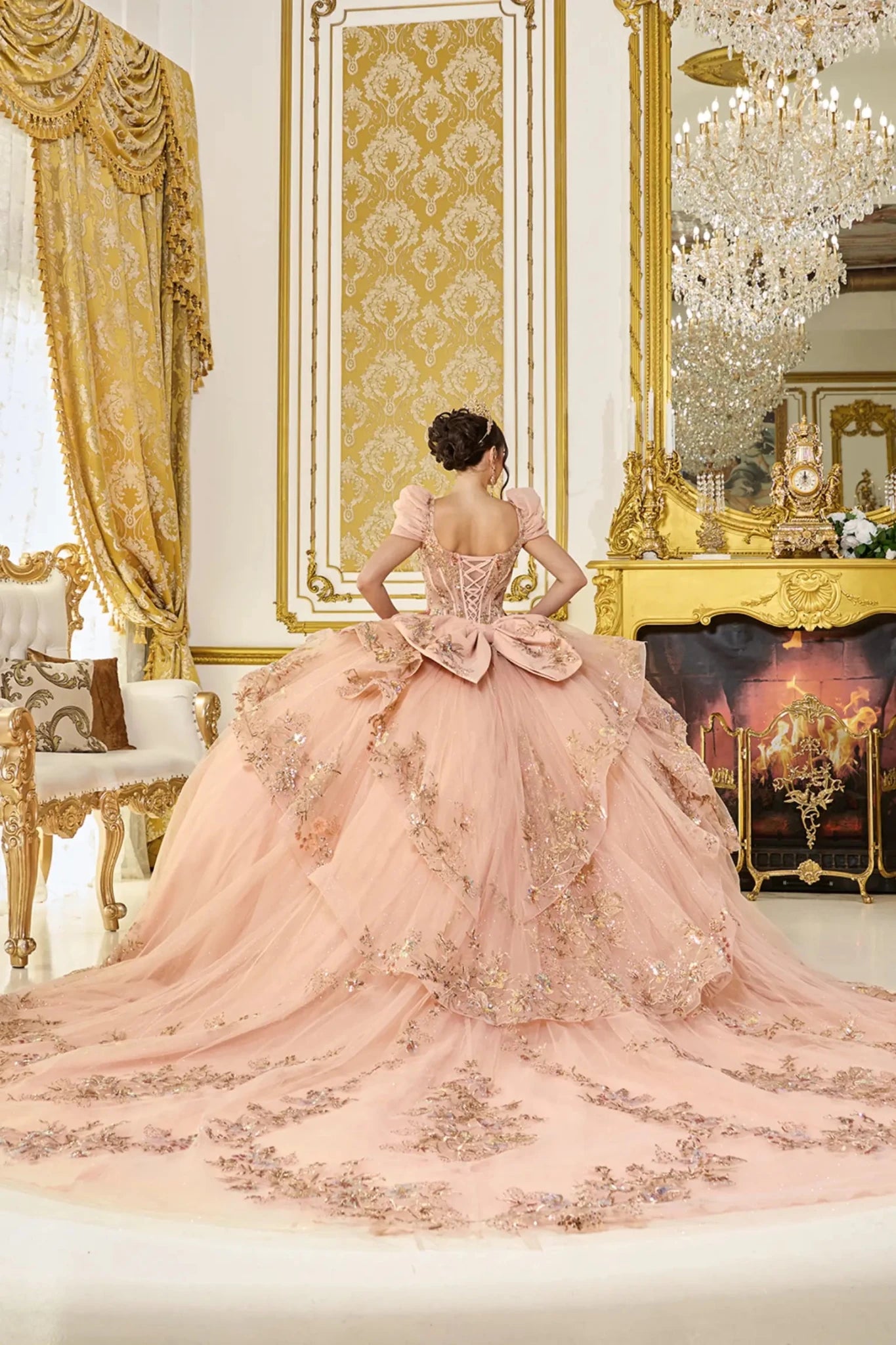 Woman standing back-facing in rose gold quinceañera ball gown with lace-up bodice, detachable cap sleeves, glitter floral appliqués, and flowing ruffled tiered skirt with train