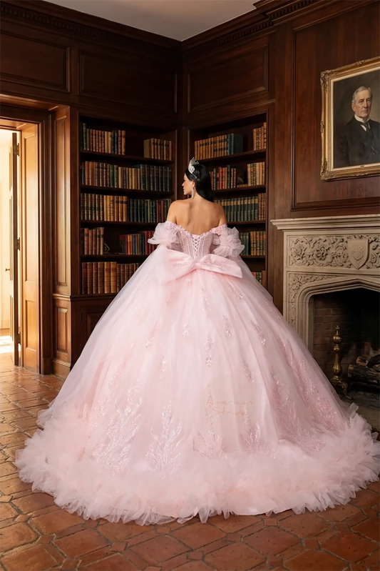 Back view of a girl wearing off‑shoulder sweetheart blush quinceañera ball gown featuring lace‑up corset closure, 3D floral appliqué, layered ruffle skirt, and detachable puff sleeves