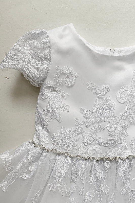 Close-up front view of a white communion dress showing lace cap sleeves, floral lace appliques, and pearl waist trim.
