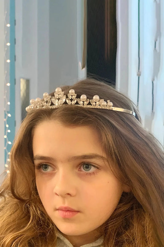 Young girl wearing a rhinestone and pearl flower motif silver tiara with a light blue curtain background