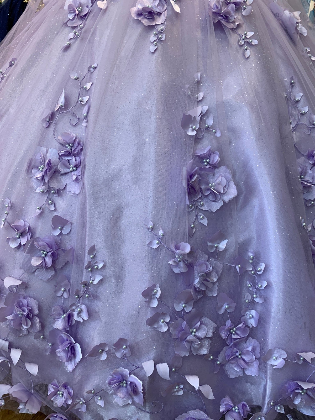 Close-up view reveals a Lilac quinceanera ball gown skirt with floral appliques, pearl embellishments, and detachable cape.