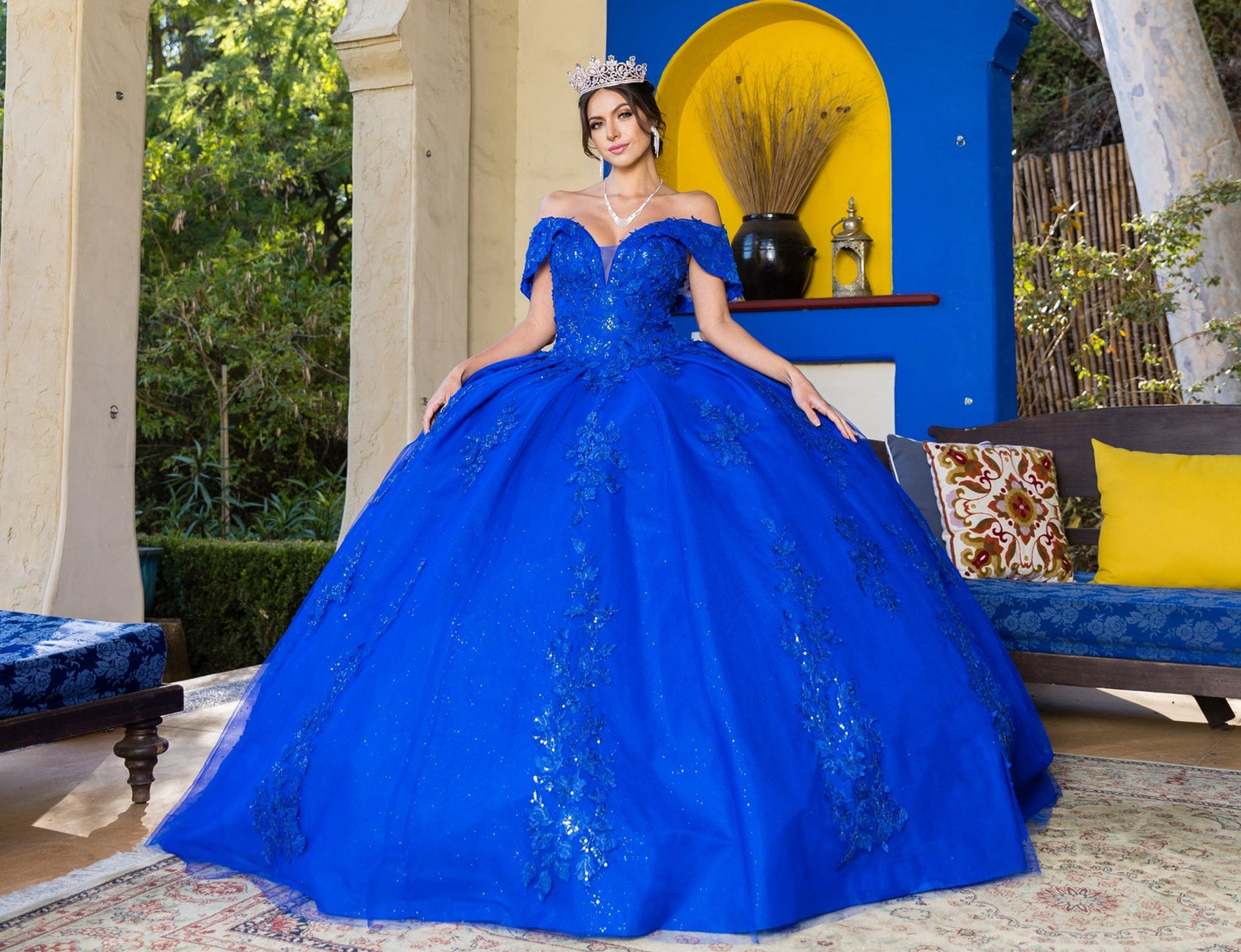 Front view of a woman wearing royal blue quinceanera dress with floral sequin appliques, glitter lace train, and layered skirt