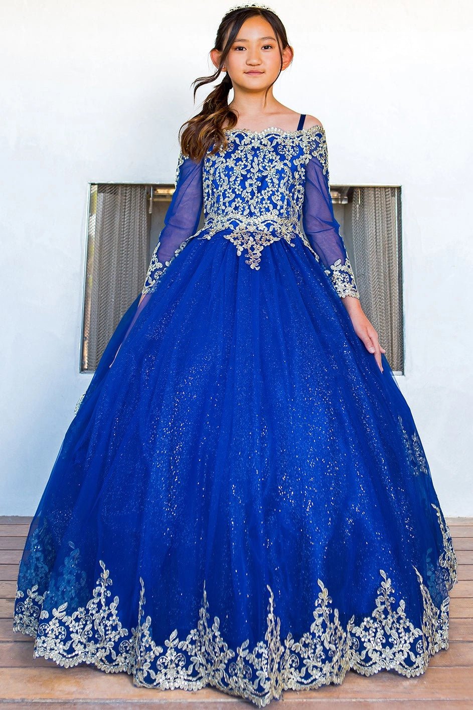 Shown in a front view, the Royal Blue mini quince dress has long sheer sleeves and gold lace over a full glitter skirt.