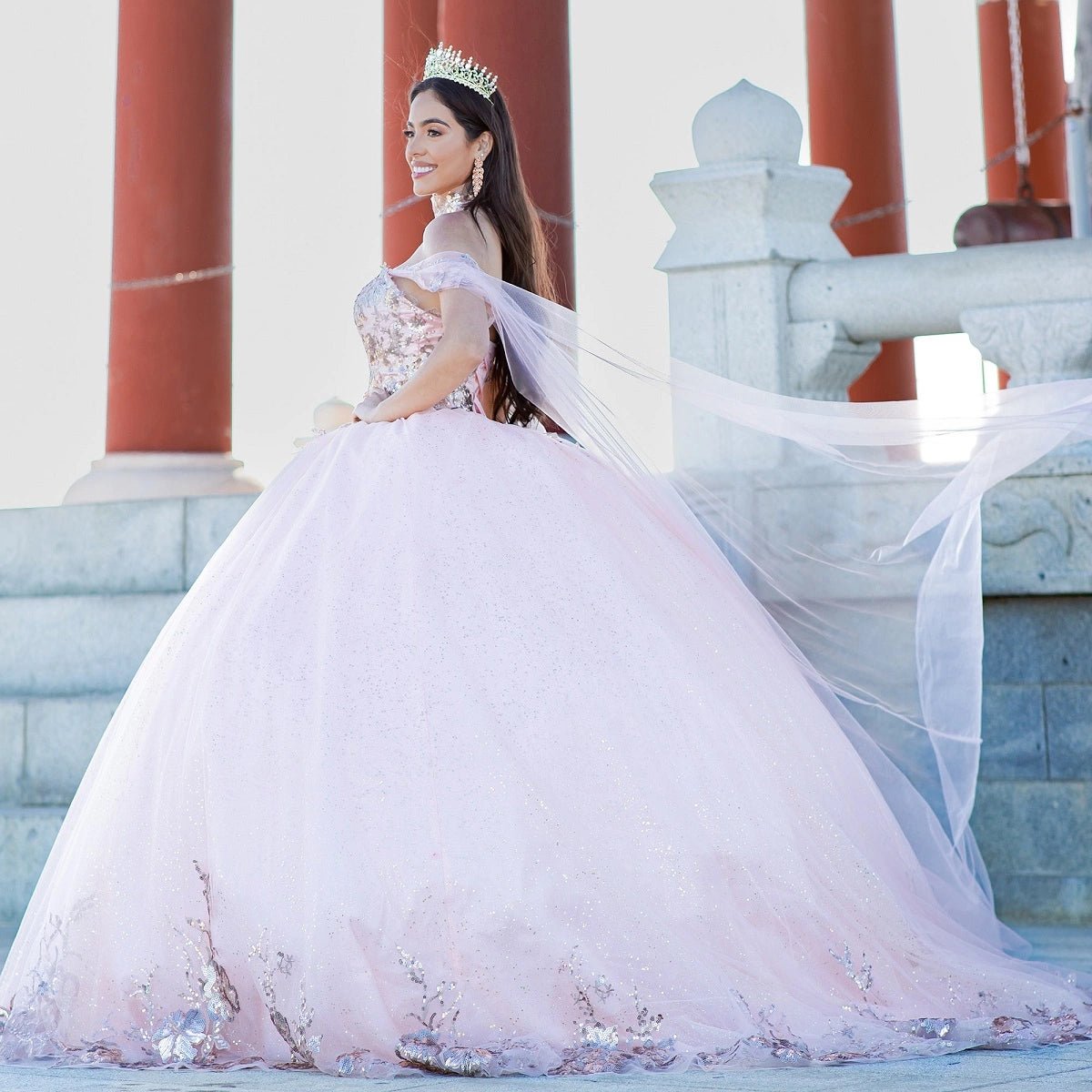 Displayed in a angled view, the Blush quinceañera dress has floral hem appliques and a sheer shoulder cape.