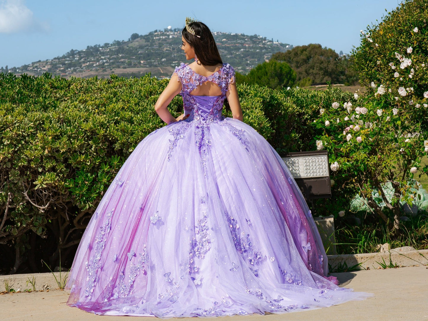 Back view of a woman wearing lilac quinceanera ball gown with embroidered lace appliques, sheer corset bodice, and ombre tulle skirt