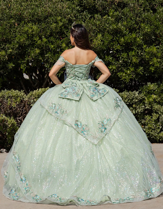 Woman in a light green ball gown with floral embroidery standing in front of green bushes.