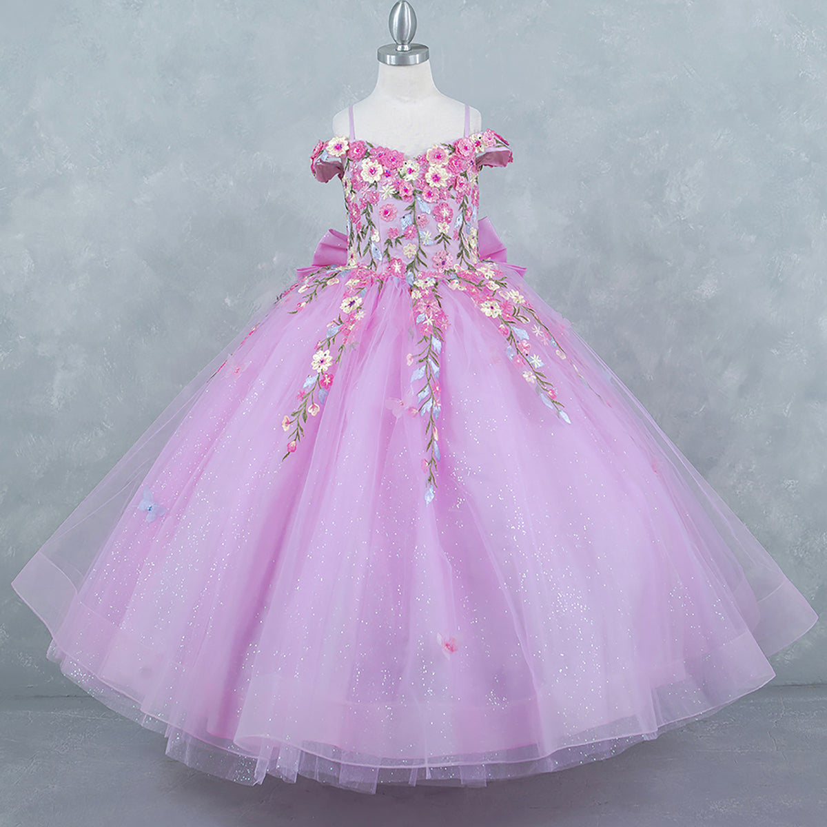 Side profile shows pink kids mini quinceañera mini quince dress with multicolor daisy embroidery and large detachable bow.