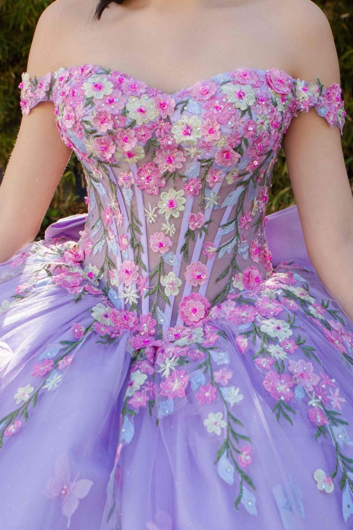 Bodice close-up highlights lilac quinceanera ball gown with multicolor daisy embroidery, detachable bow, and rhinestone detailing.