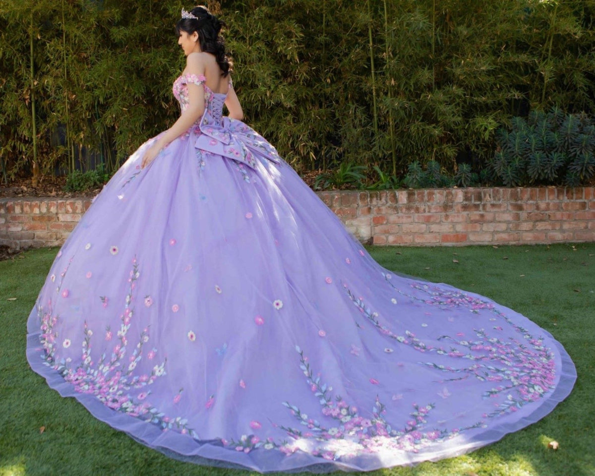Side profile shows lilac quinceanera ball gown with multicolor daisy embroidery, detachable bow, and layered skirt.