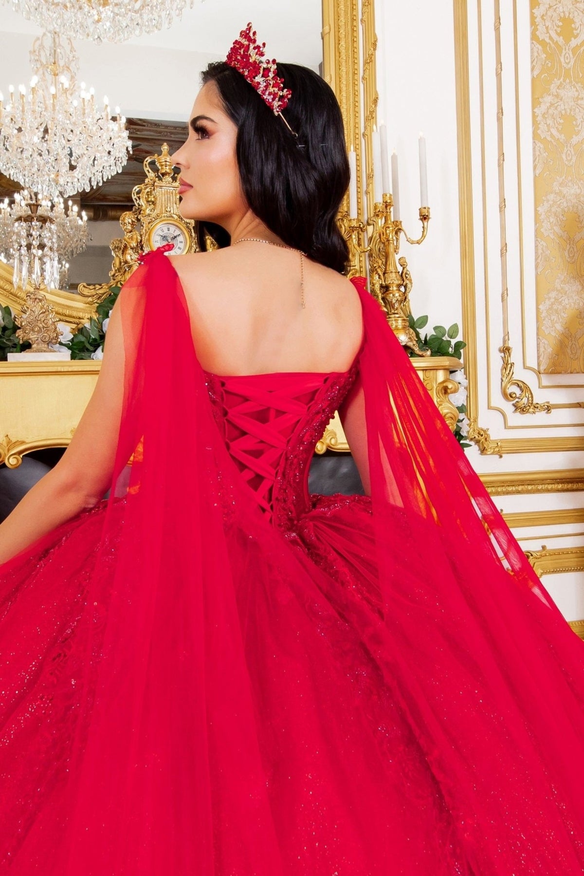 Back detail shows zoomed a red quinceanera ball gown with floral embellishments, central brooch, and flowing sheer cape.