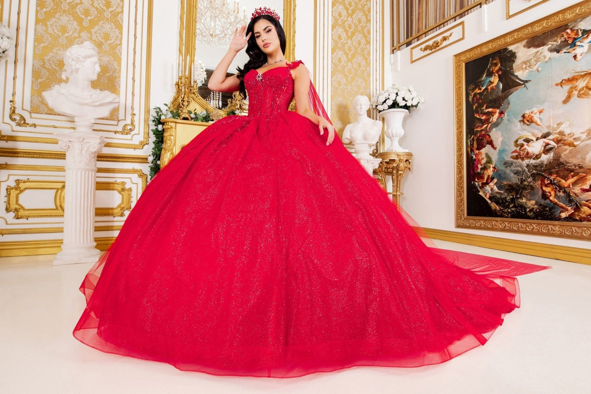 Full-length view highlights red quinceanera ball gown with floral embellishments, central brooch, and flowing sheer cape.