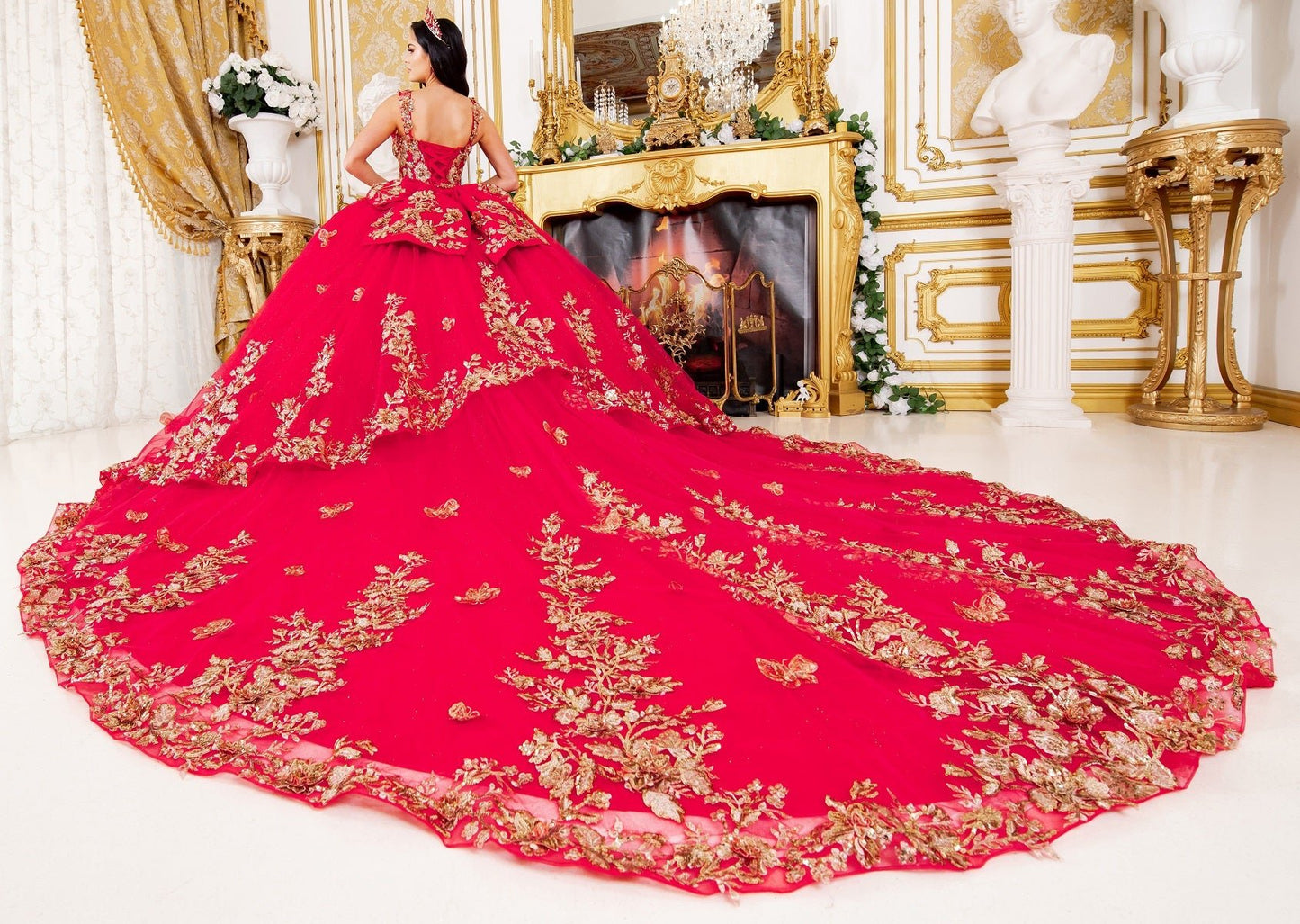 Woman standing front-facing in red ball gown with gold floral embroidery, long dramatic train, and fitted bodice in opulent indoor setting with fireplace and classical décor