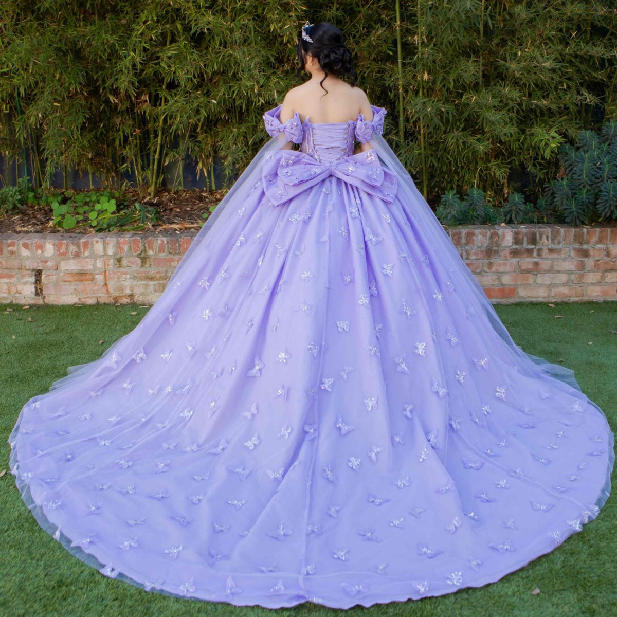 Rear view reveals lilac quinceanera ball gown with butterfly appliques, puffed sleeves, and statement bow.