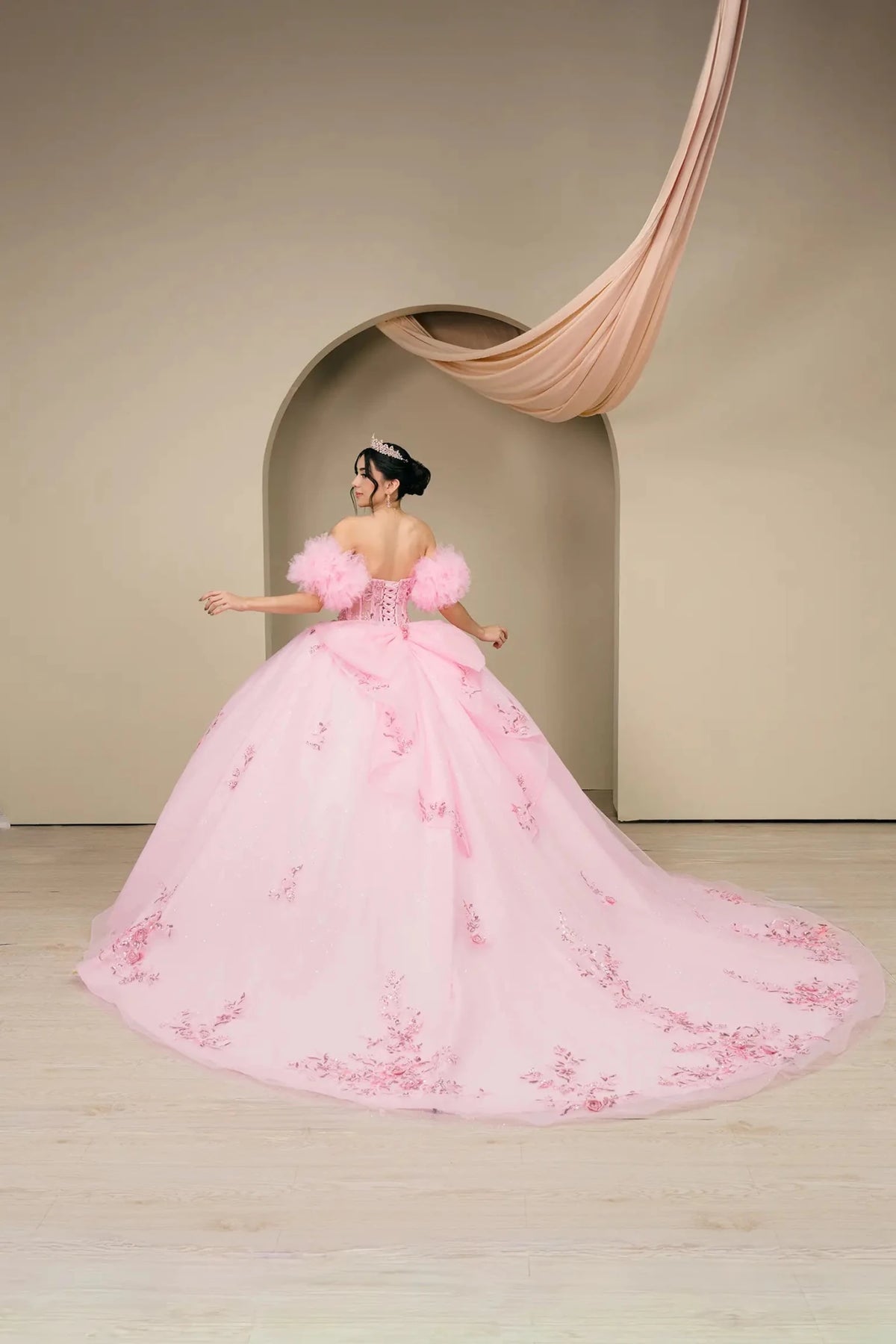 Rear view reveals a pink quinceañera ball gown showing lace-up corset, bow detail, and sweeping train.