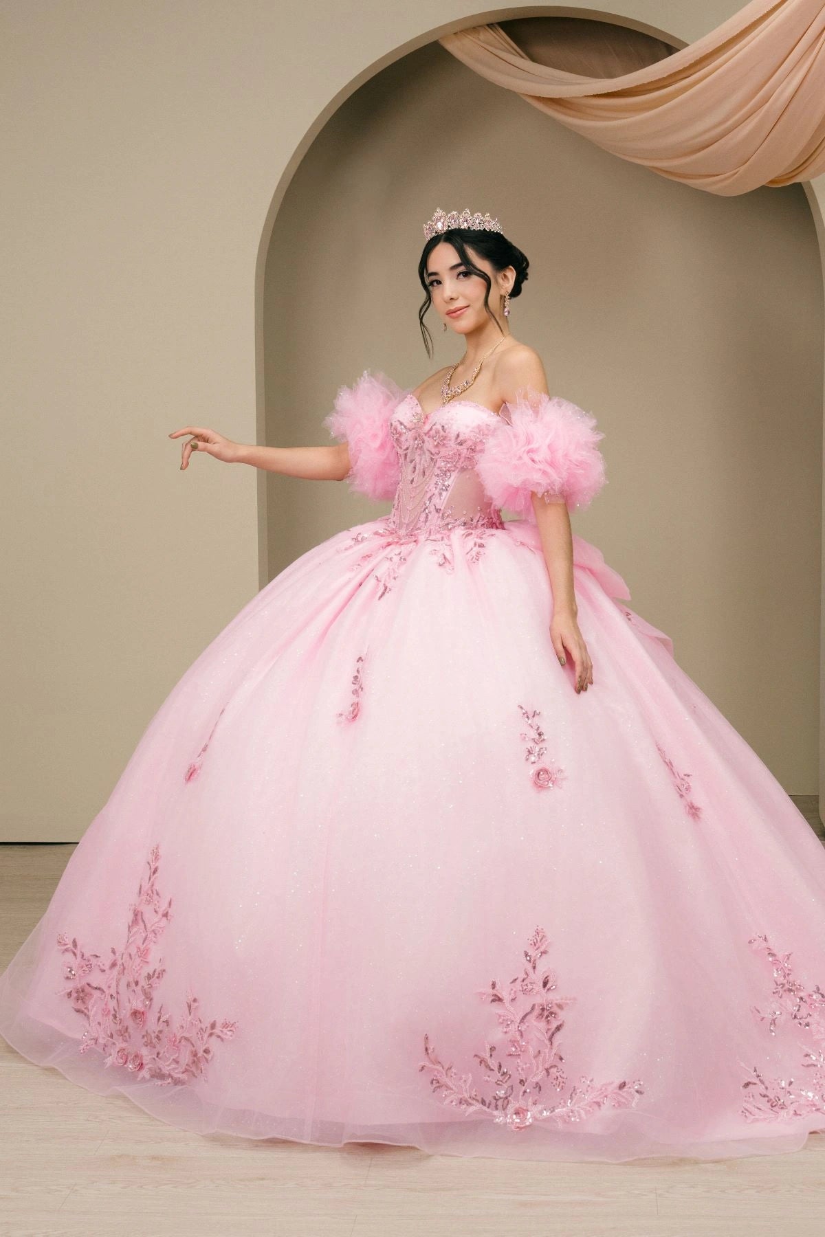 Shown in a front view, the Pink quinceañera dress has sheer puff sleeves and floral appliques across the full ball skirt.