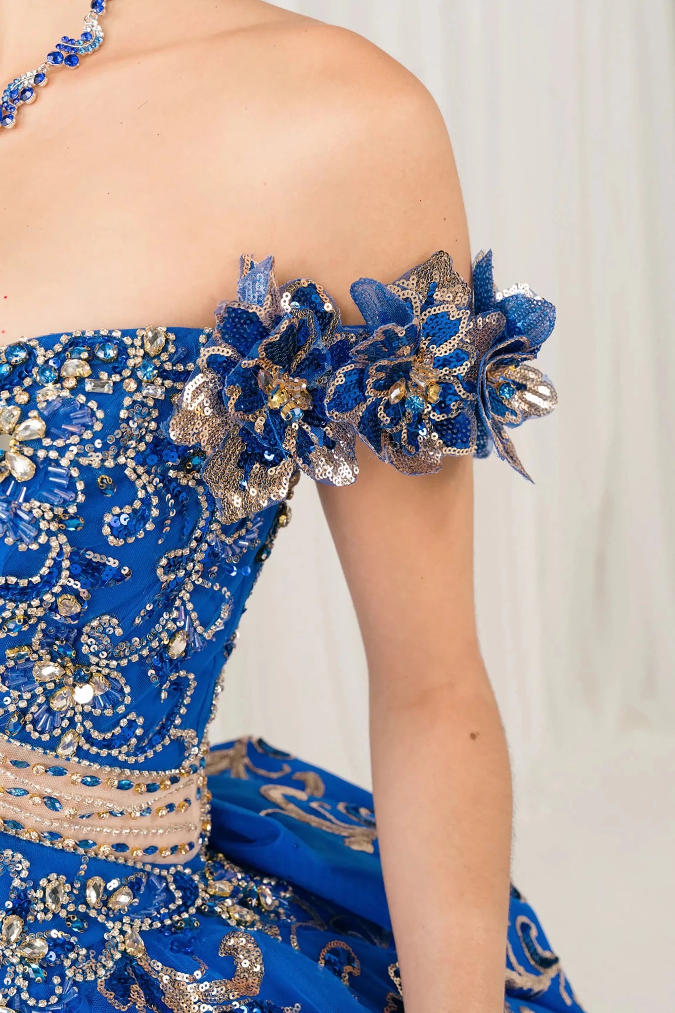 Side half close-up view of Royal quinceañera ball gown with off shoulder bodice, gold embroidery, floral appliqués, voluminous tulle skirt, corset back, and oversized bow