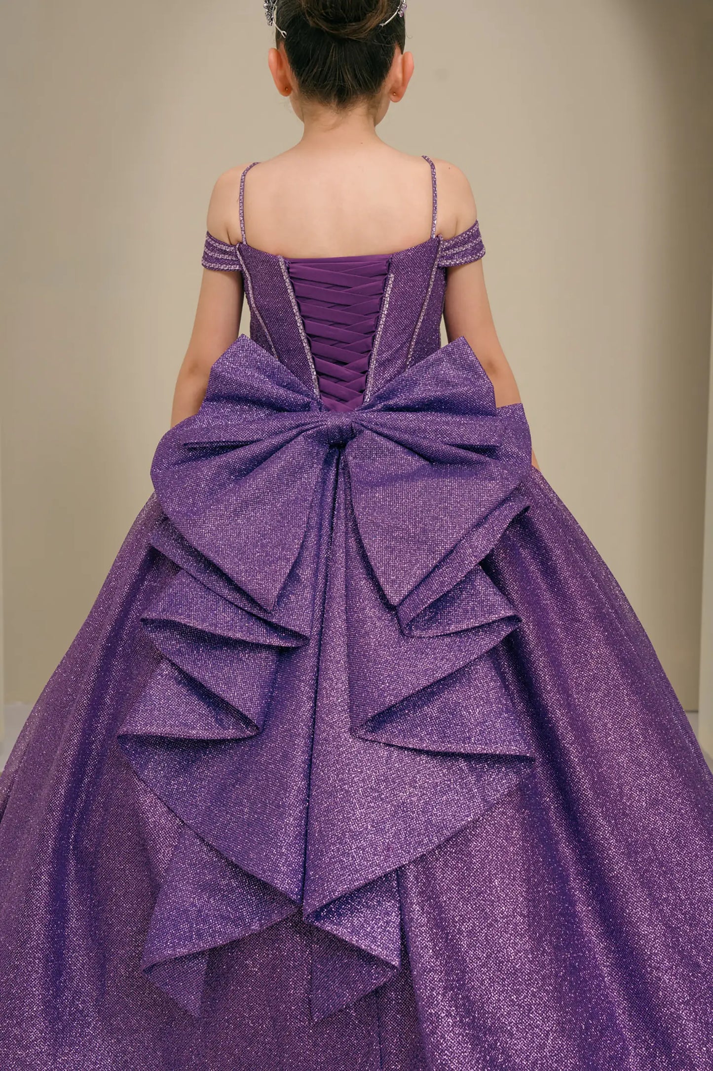 Kid standing back-facing in purple ball gown with lace-up closure, oversized tiered bow centerpiece, and cascading layered skirt