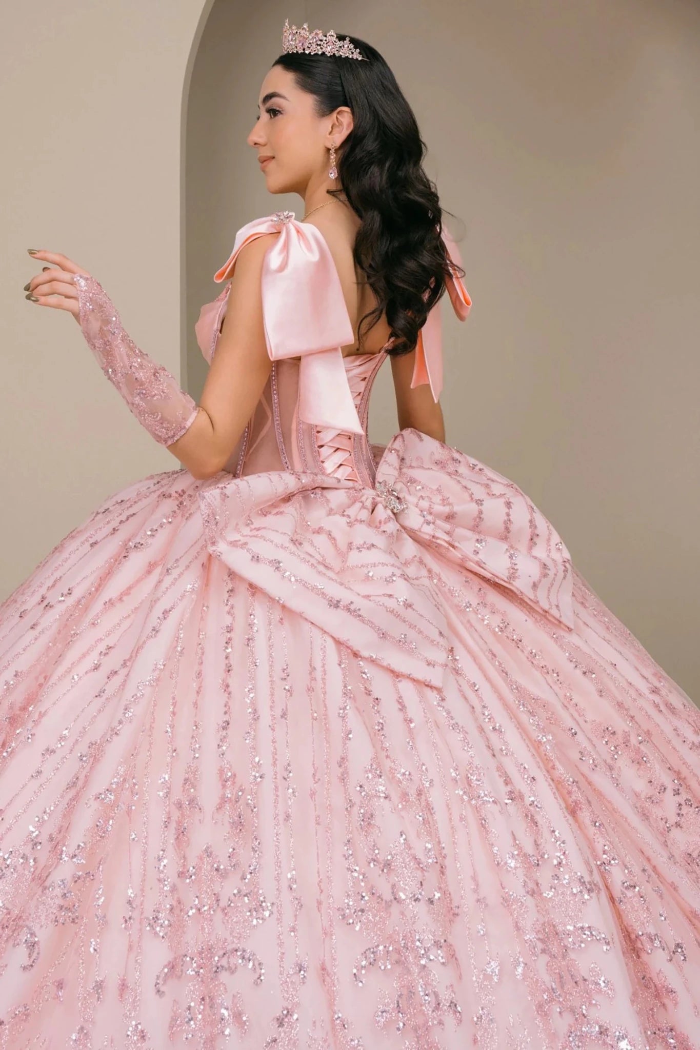 Woman wearing blush off-shoulder sweetheart quinceañera dress side-facing with beaded corset bodice, lace appliqué, layered tulle skirt, and back bow accent