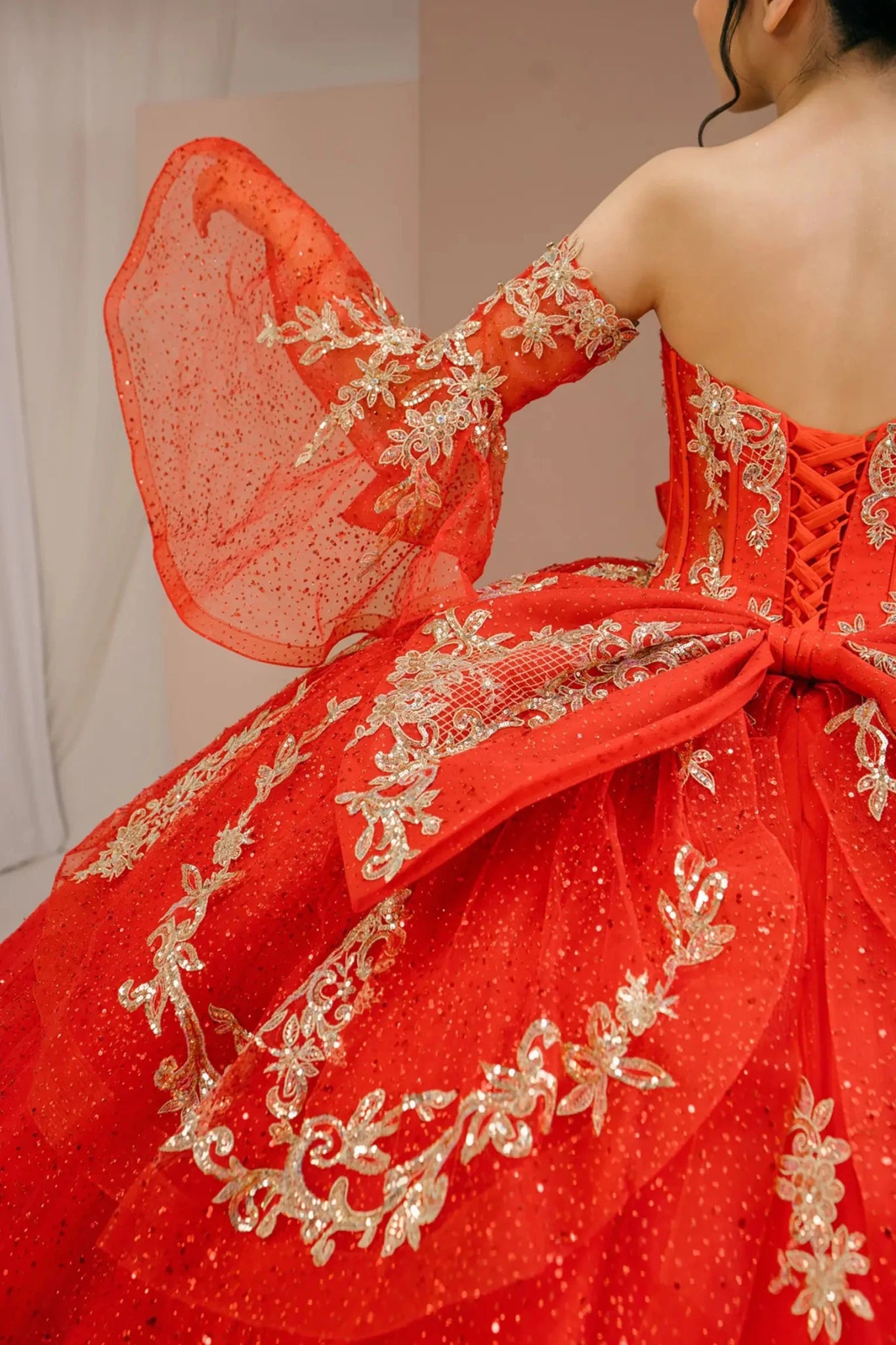 Bodice back close-up of a woman wearing red quinceañera dress with gold embroidery, sequins by flowing sleeves.