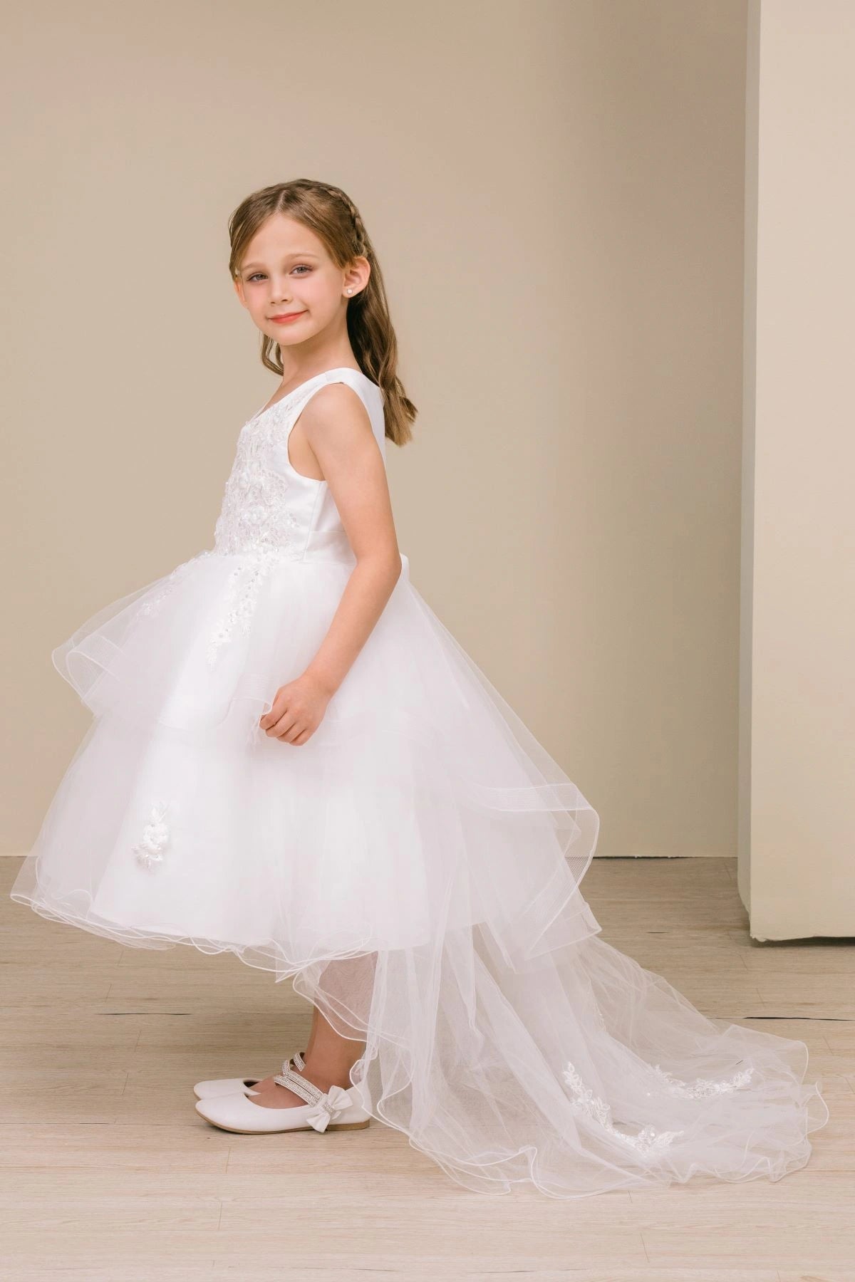 Shown in a angled view, the White flower girl dress has sleeveless bodice and tiered high-low tulle skirt with a train.