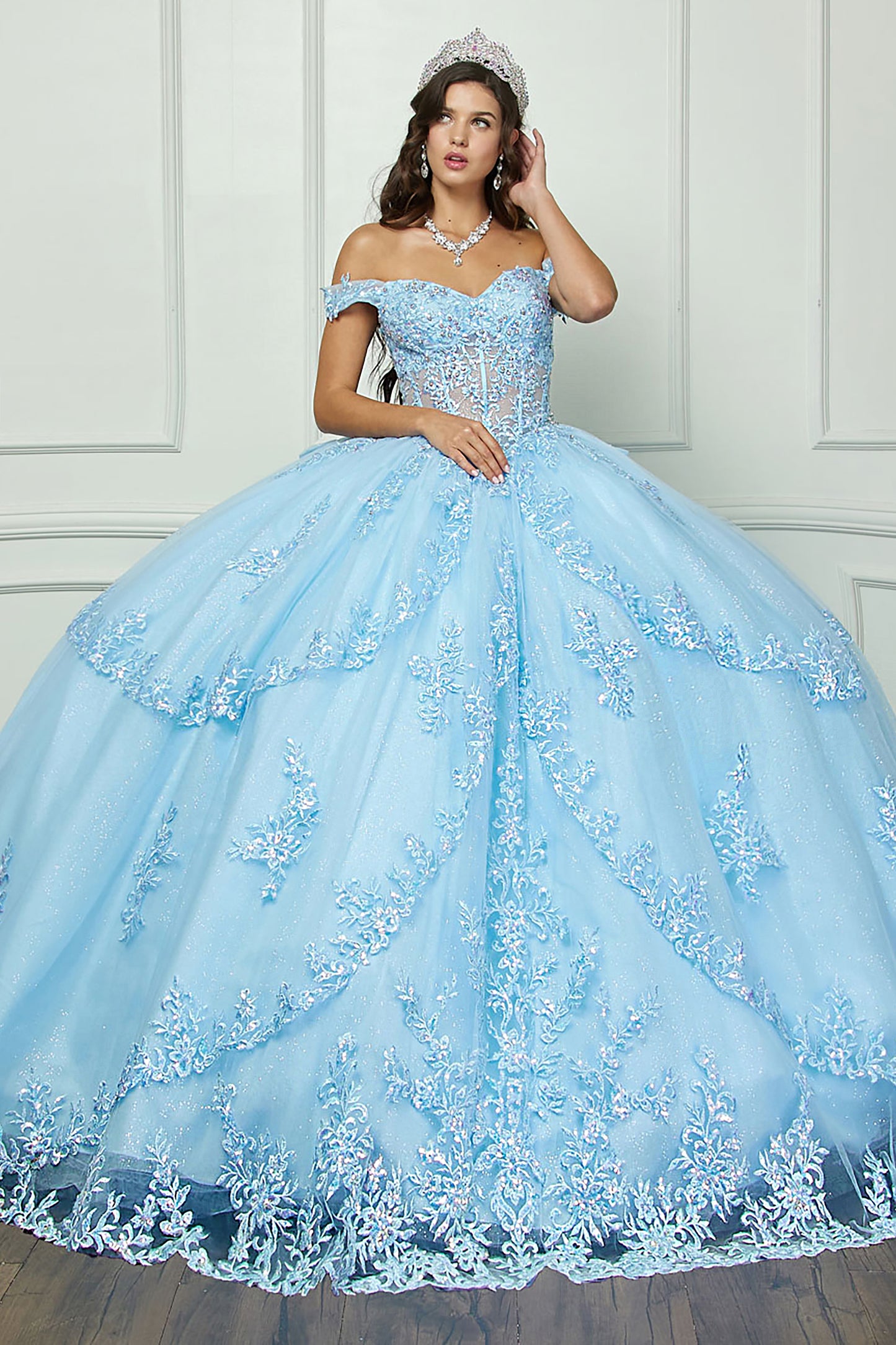 Woman wearing a light blue quinceanera dress with floral embroidery against a white paneled wall.