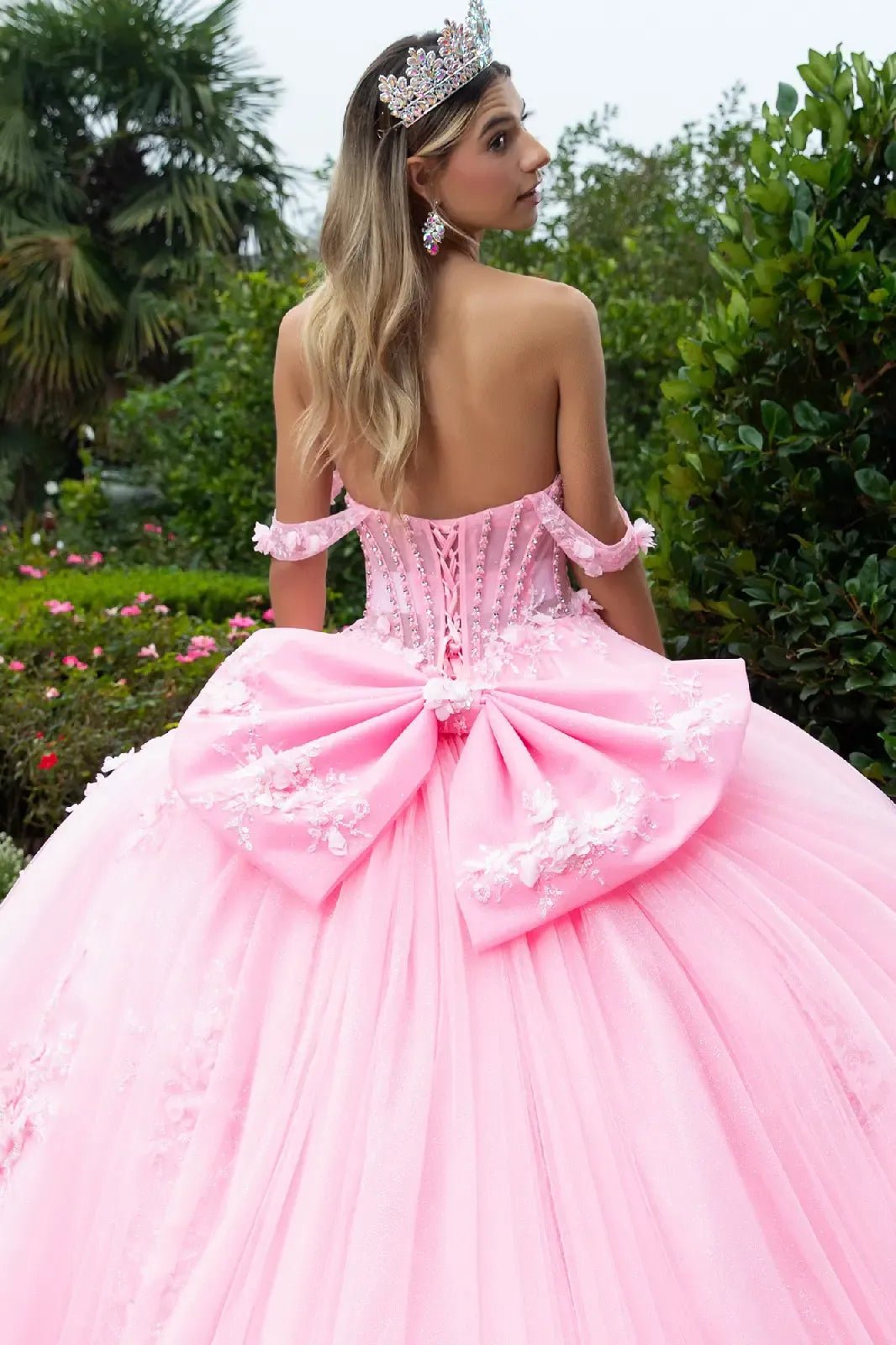 woman standing in a lush garden with palm trees and greenery. She is wearing an elaborate pink ball gown featuring a voluminous tulle skirt and a large decorative bow on the back.