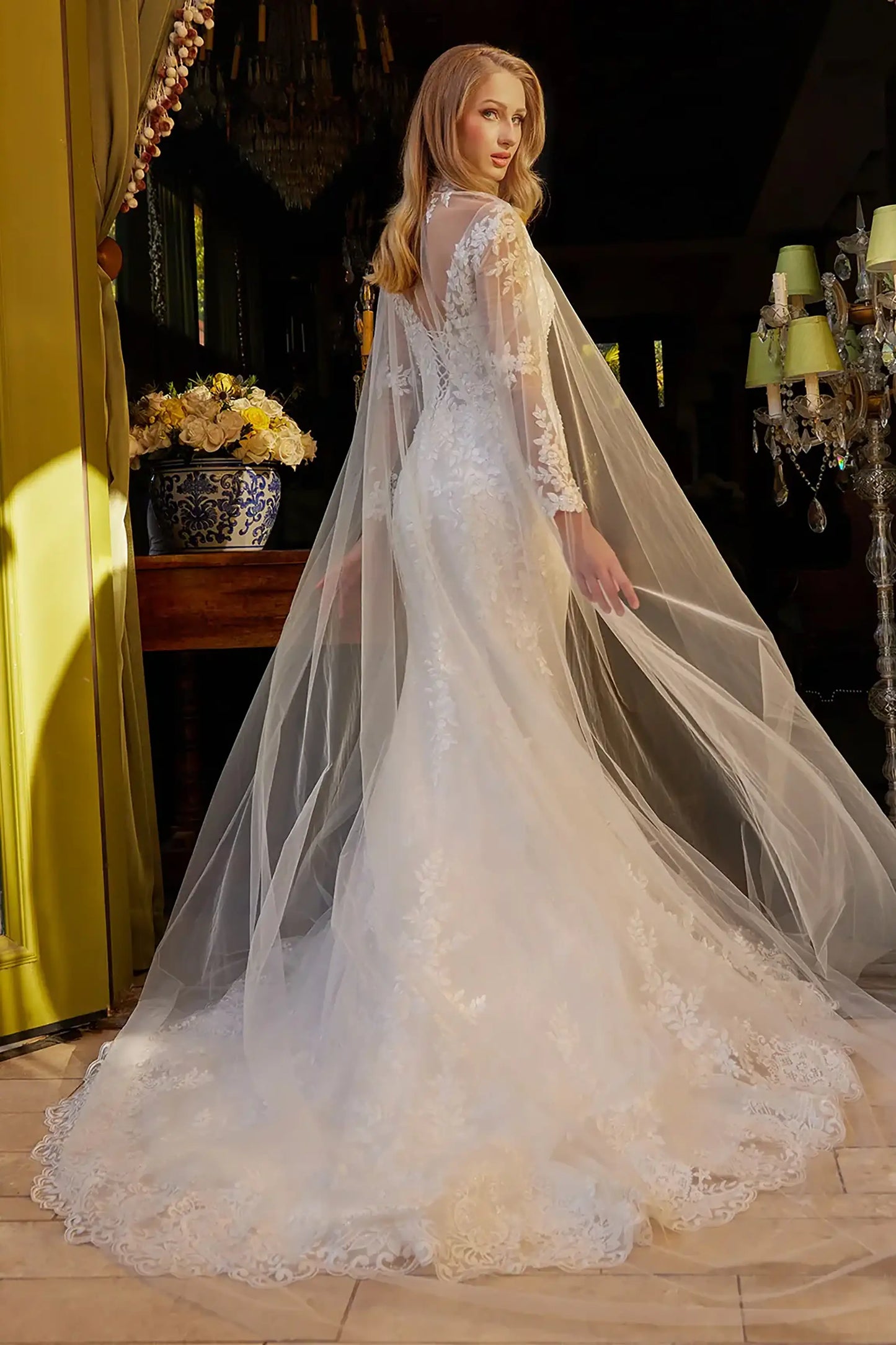 Back full-length view of Off White mermaid wedding dress with V-neck corset bodice, long sheer mesh sleeves, beaded embroidery, lace-up back, flared skirt with embroidered hem, and matching choker mesh cape
