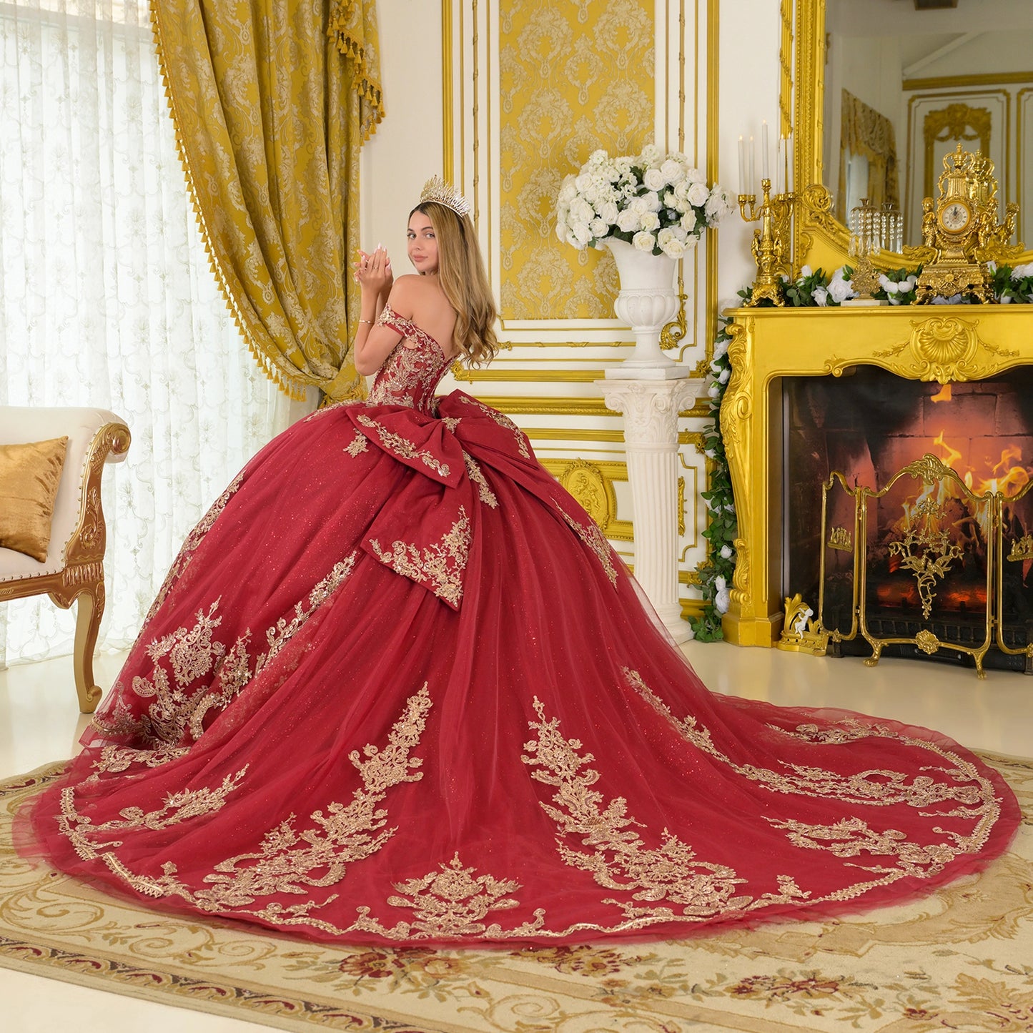 Back view of burgundy quinceañera dress with off-the-shoulder corset bodice, oversized detachable bow, gold embroidery, glitter tulle skirt, and long train