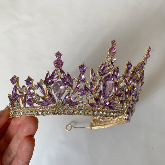 Purple and gold tiara held by a hand against a plain background