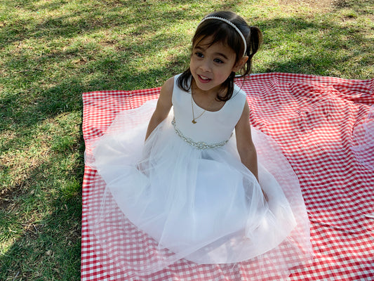 Young girl in a white dress sitting on a red and white checkered blanket in a grassy area.