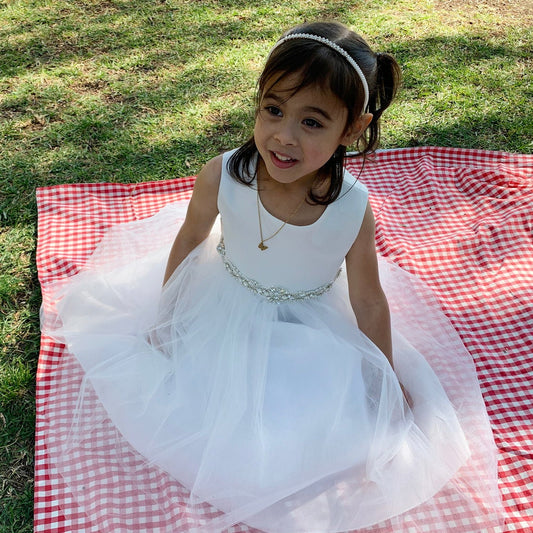 Young girl in a white dress sitting on a red and white checkered blanket in a grassy area.