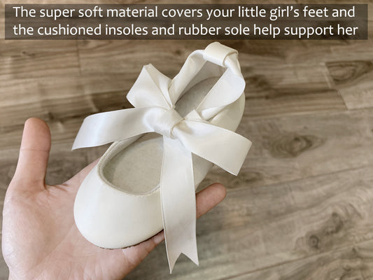 White ballerina shoe with a bow held by a hand on a wooden floor.