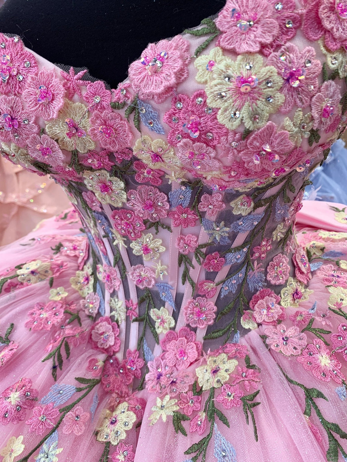 Close-up view reveals Pink quinceanera dress with pink quinceanera bodice with sheer panels, multicolor floral appliques, sequins.